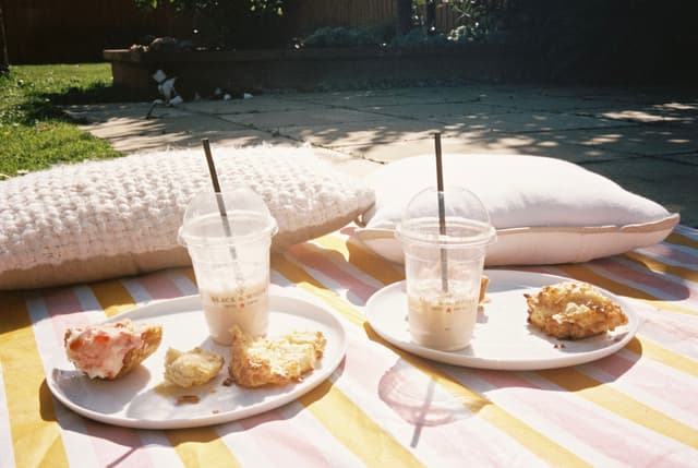 Pillow Pillows Blanket Stripes Plates Plate Pastries Pastry Muffin Muffins Drink Drinks Cups Cup Straw Straws Outdoor Outdoors Grass Lawn Sunlight Shadows White Yellow Pink Food Beverage Picnic Relaxation Summer