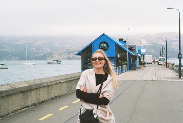 woman sunglasses smiling blue building water boats pier road lamp post mountains cloudy sky jacket bag pathway railing coast