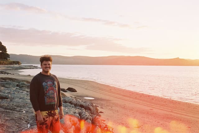 Beach Ocean Water Sand Person Sunset Sky Clouds Rocks Shoreline Landscape Horizon Light Flare Trees Coastline Casual Clothing Outdoors Nature Scenic Calm Tranquil Relaxation Leisure Daytime Warmth Reflection Travel Adventure Primary Color: Orange