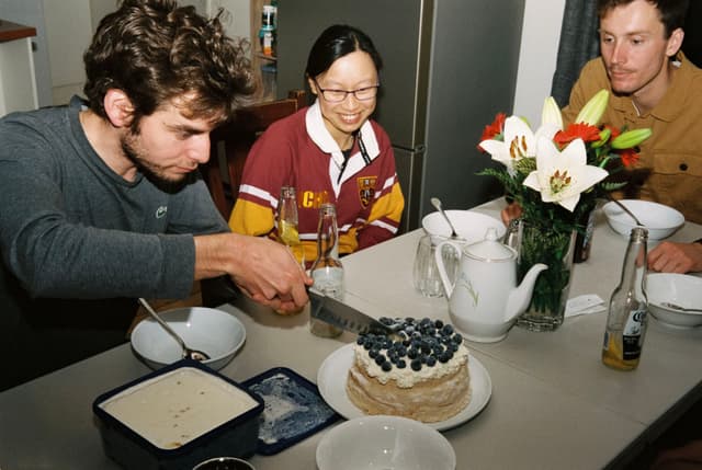 cake blueberries flowers people table plates bowls knife teapot drinks bottles ice cream dessert dining room celebration gathering meal white red green brown black gray cutting serving eating smiling sitting vase leaves stem petals beverage glass container spoon fork napkin casual socializing primary color white