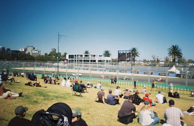race track cars spectators grass sky fence buildings trees palm trees stroller blue