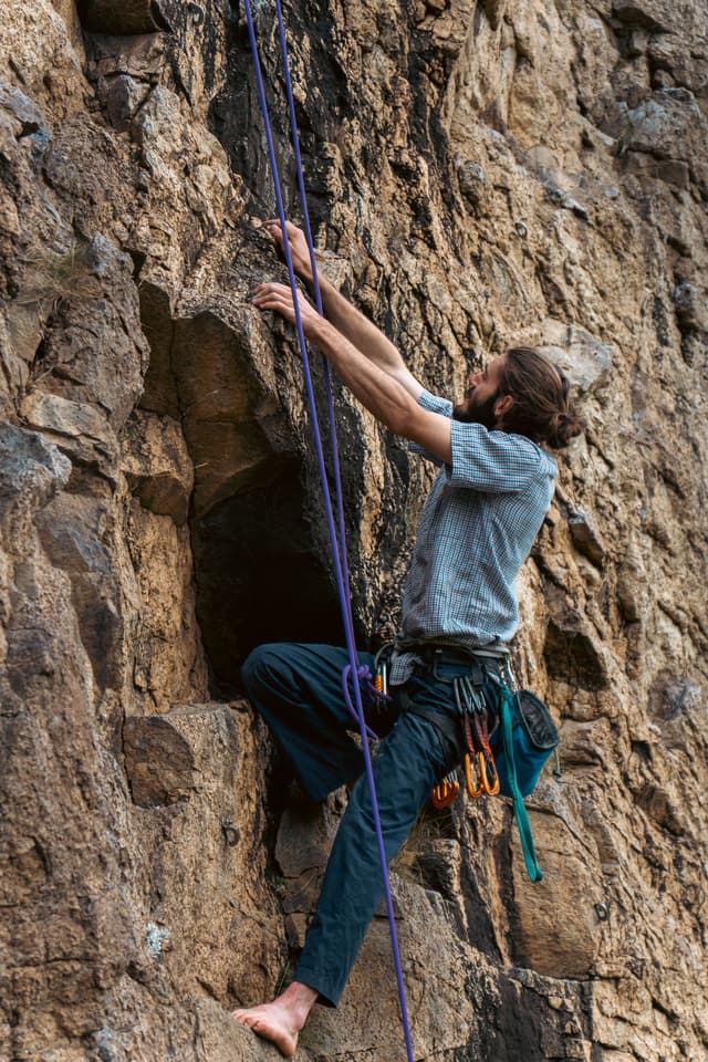 Climbing Rock Climber Rope Wall Gear Harness Shoes Outdoors Adventure Sport Nature Brown Blue Gray Rugged Cliff Vertical Ascent Challenge Strength Focus Determination Safety Equipment Physical Activity Effort Concentration Movement Skill