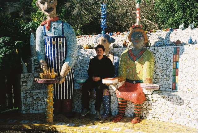 sculpture statue mosaic art garden outdoor colorful human figure sitting standing striped apron yellow red blue green trees bushes wall bench stone gravel