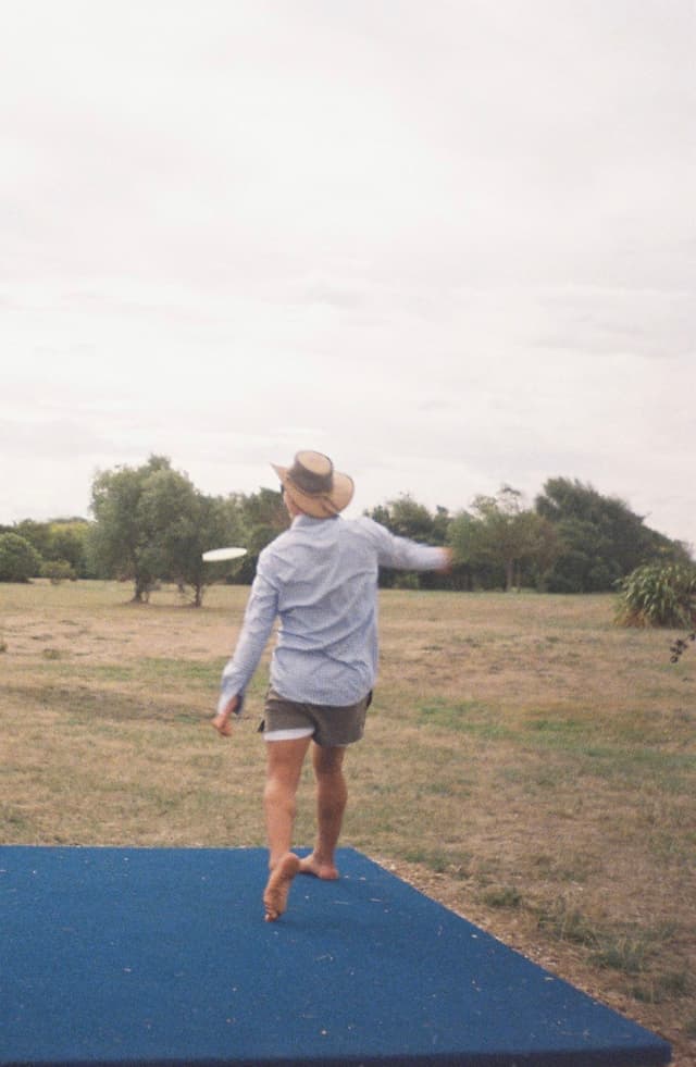 person hat shirt shorts grass field trees sky outdoors barefoot blue standing walking nature landscape casual summer
