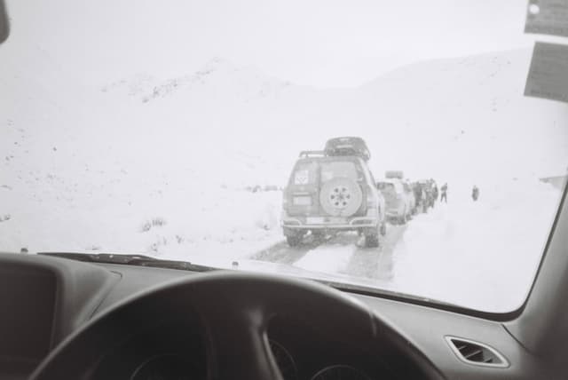 Car SUV Snow Road Steering Wheel Windshield Mountains Traffic Winter White Cold Journey Travel Outdoors Landscape Overcast Weather Adventure Drive Vehicle Queue Rearview Mirror Tires Roof Rack Expedition Pathway Horizon Overhead Cloudy Rugged Terrain