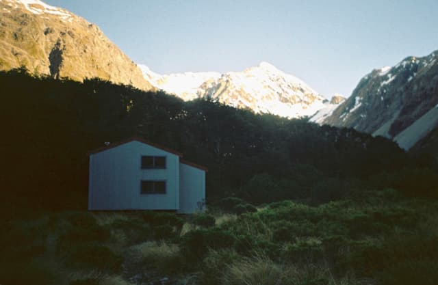 House Cabin Mountains Snow Trees Forest Landscape Nature Sky Blue Green Shadows Sunlight Hills Valleys Peaks Roof Windows Walls Grass Bushes Rocks Cliffs Scenery Outdoors Remote Tranquil Peaceful Quiet