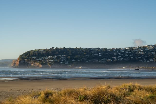 Beach Ocean Water Waves Sand Grass Hill Houses Sky Blue Coastline Shoreline Landscape Nature Scenery Horizon Cliff Seaside Sea View Dunes Vegetation Morning Calm Tranquil Peaceful Coastal Residential Summer