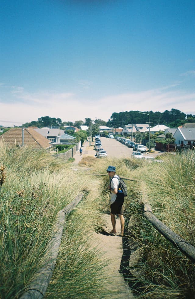 path grass person hat backpack houses road cars sky blue trees sand fence walkway summer outdoor nature landscape