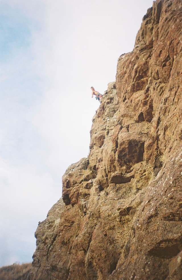 rock cliff climbing person sky clouds brown blue rugged outdoor adventure nature vertical height landscape formation texture ruggedness edge summit rocky surface elevation exploration challenge ascent precipice terrain geological feature cliffside craggy steep ruggedness outdoorsman daring verticality
