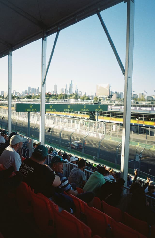 stadium race track spectators grandstand city skyline buildings sky blue seats people event outdoor architecture railing urban sports daytime