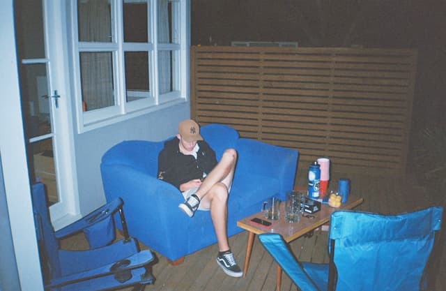 A person wearing a cap is sitting on a blue couch on a patio, surrounded by chairs and a table with various items on it