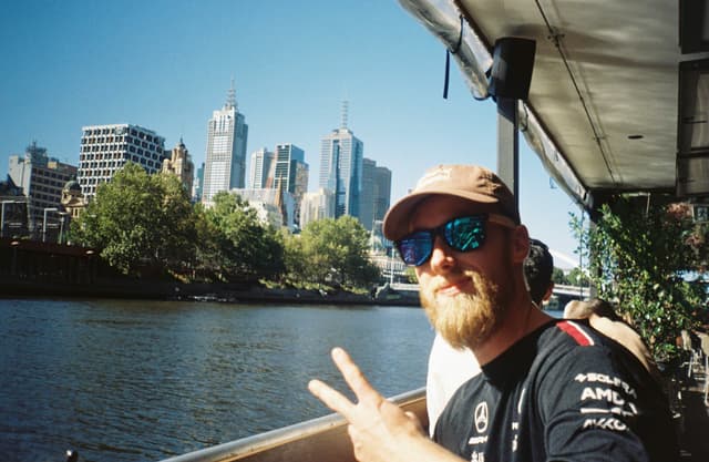 man sunglasses beard cap peace sign cityscape buildings river water trees sky blue outdoor urban bridge railing reflection daytime