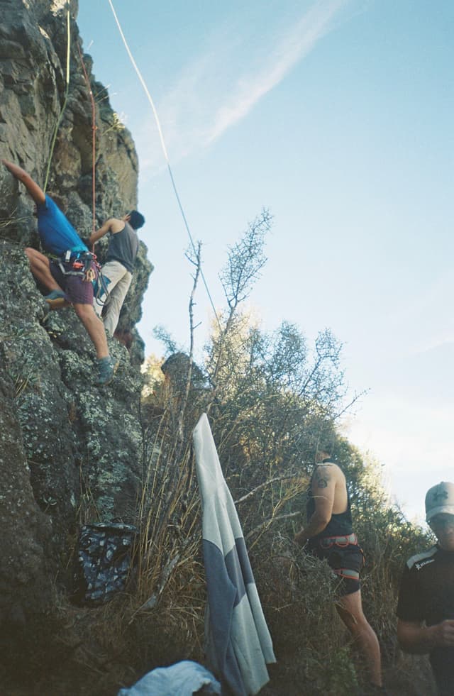 Climbing Rock Climber Rope Harness Helmet Outdoor Nature Sky Blue Cliff Vegetation Bushes Gear Equipment Adventure Sport Activity Challenge Vertical Ascent Safety Protection Teamwork Group Effort Exercise Fitness Rugged Terrain Exploration Courage Determination