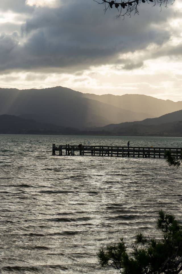 lake water pier dock mountains hills clouds sky sunset silhouette landscape nature outdoor scenery reflection horizon tree branches gray