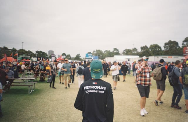 people crowd festival outdoor grass trees sky tents tables chairs walking standing blue black green red white gray backpack hat jacket shirt shorts pants sneakers