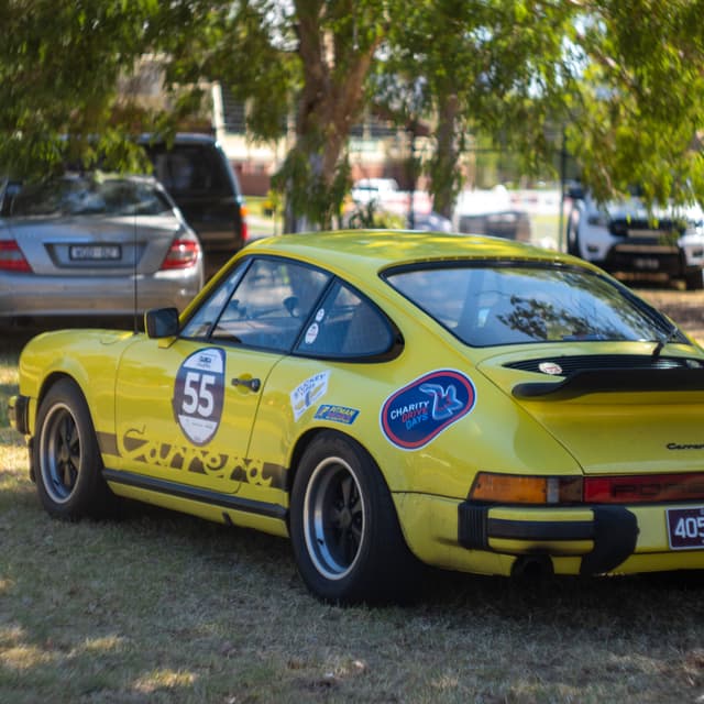 Yellow car vehicle racecar number 55 stickers trees grass park rear view wheels tires windows bumper license plate sunlight shadow outdoors classic vintage sports car parking lot nature automotive transportation decals competition retro sporty shiny summer day environment landscape background