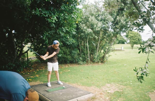 golf player swing club trees grass outdoors green shorts cap shirt shoes tee nature foliage bushes leaves pathway standing action summer sports recreation leisure activity