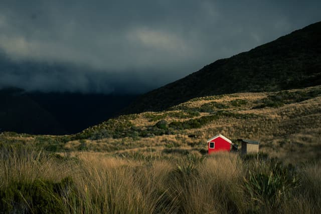 landscape hills grass hut cabin red dark sky clouds nature outdoors rural scenery field vegetation mountain moody overcast stormy wilderness solitude remote isolated countryside natural environment terrain shadow dramatic peaceful tranquil serene atmospheric rugged wild untouched primary color red