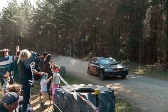 car race rally dirt road spectators trees forest dust motion sunlight blue green red black white gray people children adults fence cheering action speed excitement outdoors daytime