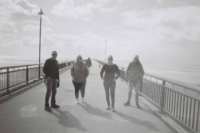 people bridge railing sky clouds lamps walkway black white grayscale shadows ocean horizon pathway
