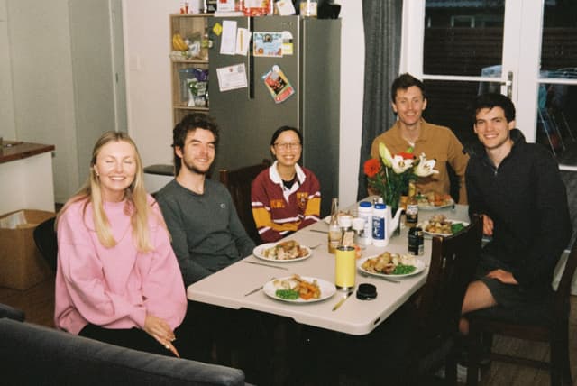 People Table Dinner Food Plates Glasses Flowers Refrigerator Photos Smiles Chairs Window Door Pink Shirt Gray Shirt Red Shirt Brown Shirt Black Shirt Yellow Cup White Walls Kitchen Dining Room Meal Gathering Group Celebration Home Interior Primary Color: Pink