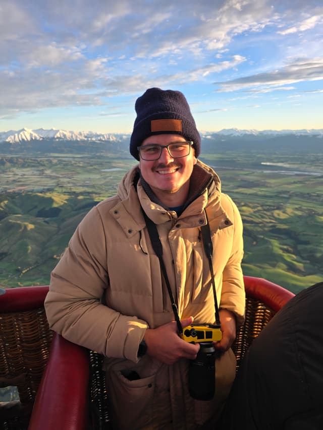 hot air balloon basket person coat beanie hat glasses camera landscape sky clouds mountains snow green fields horizon sunrise morning outdoor adventure travel photography smile red blue beige black yellow