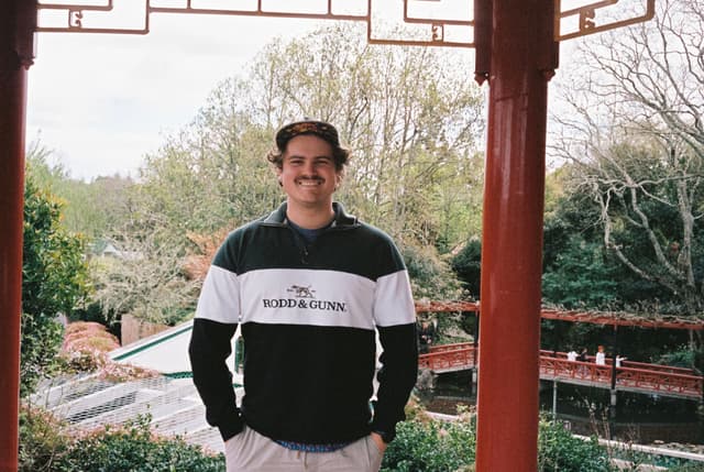 person man smiling standing outdoors trees greenery garden pavilion red columns roof sky casual clothing hat mustache nature architecture landscape background spring foliage leisure portrait photography travel tourism relaxation peaceful serene primary color red