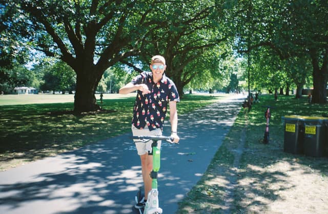 park trees path person scooter shirt shorts sunlight shadow grass green summer outdoors riding leisure nature transportation fun casual day foliage recreation electric scooter