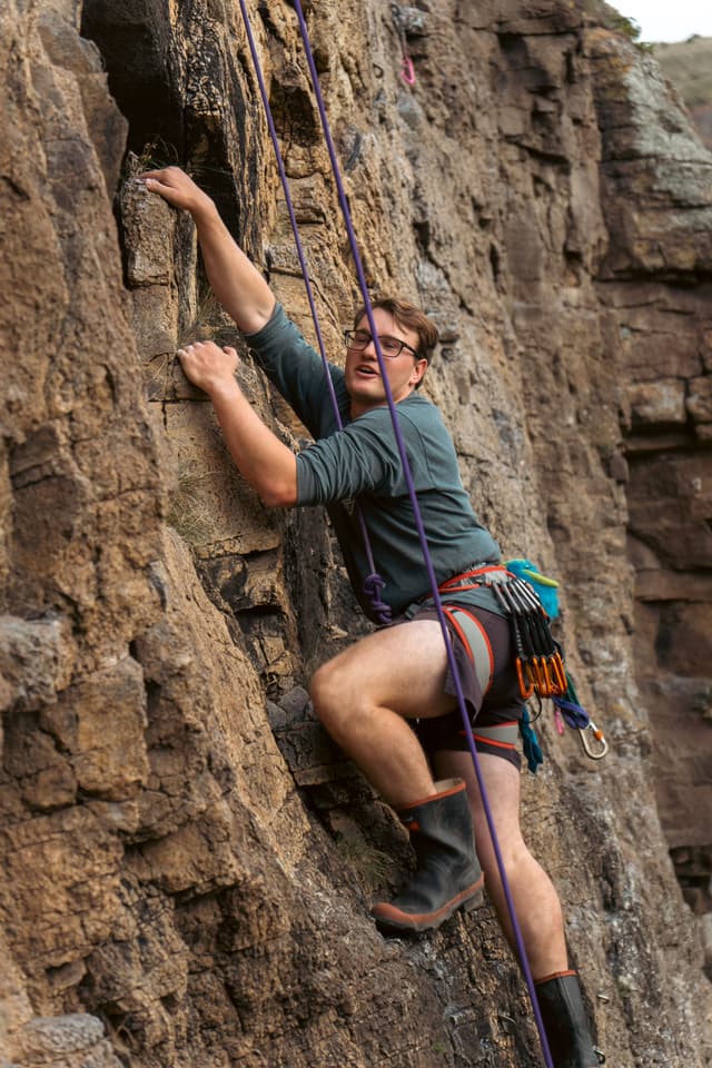 rock climbing climber rope harness helmet rock wall cliff outdoor adventure sport safety gear boots brown green blue gray rugged ascent challenge vertical nature equipment protection concentration effort determination focus strength balance technique exploration physical activity recreation primary color brown