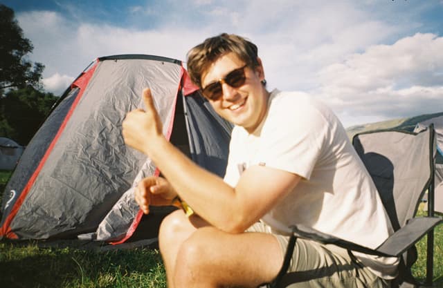 Man Tent Sunglasses Chair Grass Sky Cloud Thumbs Up Bottle Camping Outdoor Daylight Casual Relaxation Smiling Summer Leisure Nature Travel Adventure Recreation Happy Fun Shade Sunlight Blue Green Red White Black