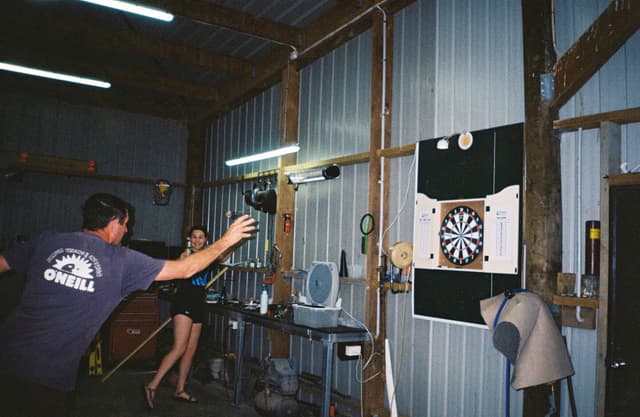 dartboard darts game throw player person barn wall light wood metal blue shirt shorts mannequin table chair