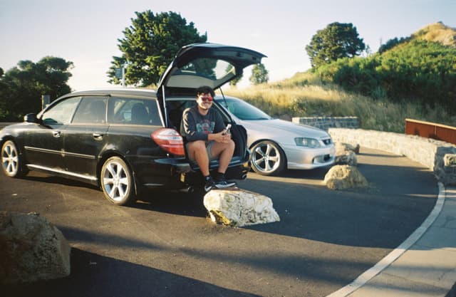 car vehicle person sitting road rock tree grass sky outdoor black white silver blue green stone hill landscape parking sunlight shadow casual relaxed summer travel journey transportation nature scenery
