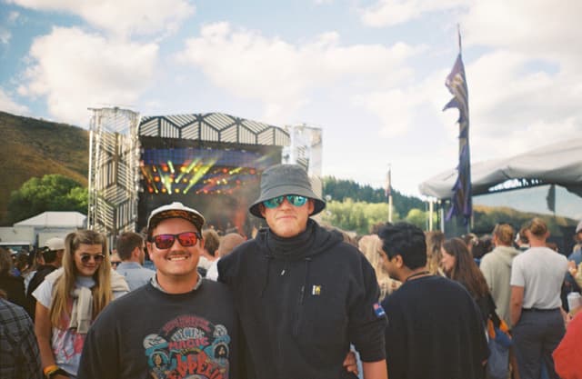 Two people wearing sunglasses and hats stand in front of a crowd at an outdoor music festival, with a stage and hills in the background