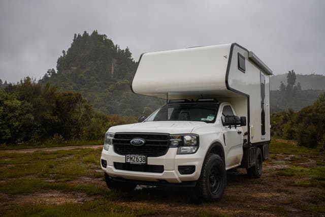 camper van truck vehicle white overcast sky grass trees forest mountain landscape outdoors travel adventure camping road trip nature exploration rugged wilderness journey transportation scenery cloudy
