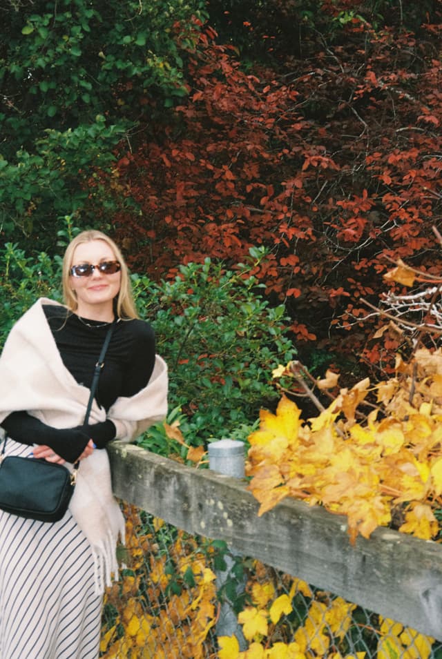 woman sunglasses scarf bag fence leaves autumn foliage trees plants yellow red green black white beige nature outdoors casual fashion smiling shawl skirt railing fall season