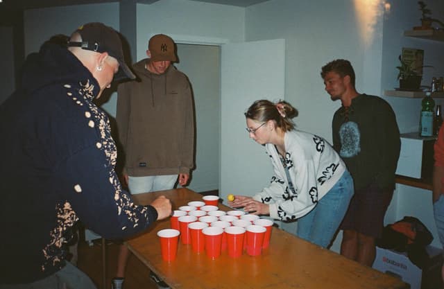 people table cups red game party indoors hoodie sweater hat bottle shelf wall light wood fun activity gathering casual standing playing interaction concentration teamwork enjoyment socializing friends competition leisure event focus strategy skill challenge entertainment beverage coordination balance aiming reflexes excitement laughter camaraderie