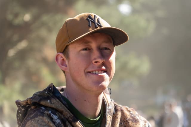 man cap hat brown jacket outdoors trees sunlight smile nature casual relaxed daytime blurred background