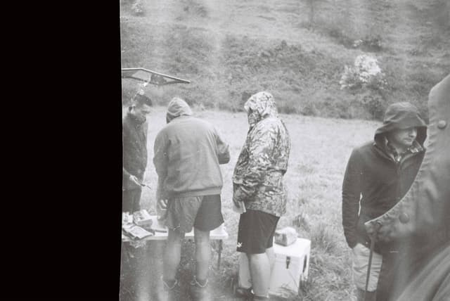 black white people outdoors coats hoods grass field table gathering overcast jackets boots nature landscape group standing rain casual clothing countryside rural scene picnic socializing weather dreary monochrome vintage retro atmosphere