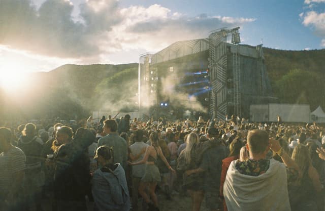 concert festival crowd stage music outdoor people audience performance event sunlight sky clouds mountains trees blue green smoke atmosphere summer celebration entertainment sound system speakers excitement gathering fun enjoyment party energy live show festivalgoers atmosphere landscape scenery