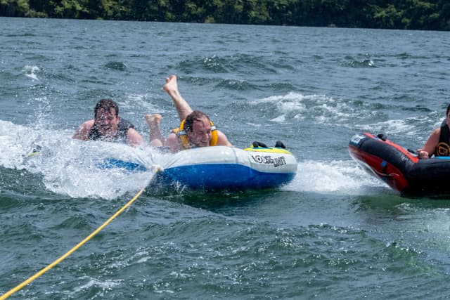 water tubing lake people inflatable tube rope splash waves trees blue yellow red black fun summer outdoor recreation adventure action speed excitement nature sports leisure activity thrill riding boating aquatic landscape forest shoreline