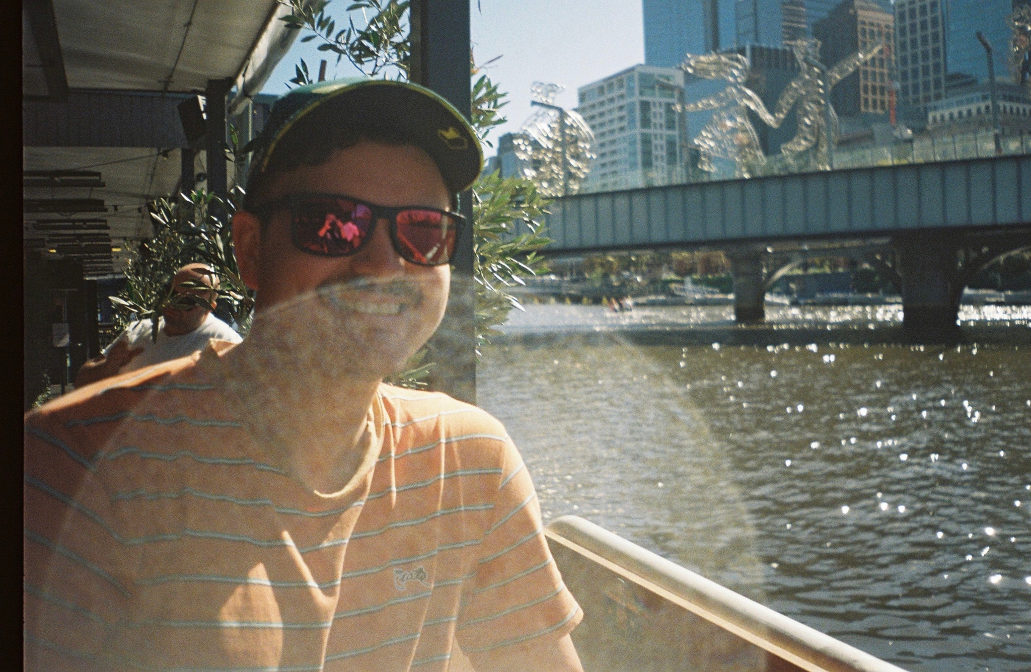 person sunglasses hat river bridge city buildings trees sunlight reflection outdoor urban water blue sky railing