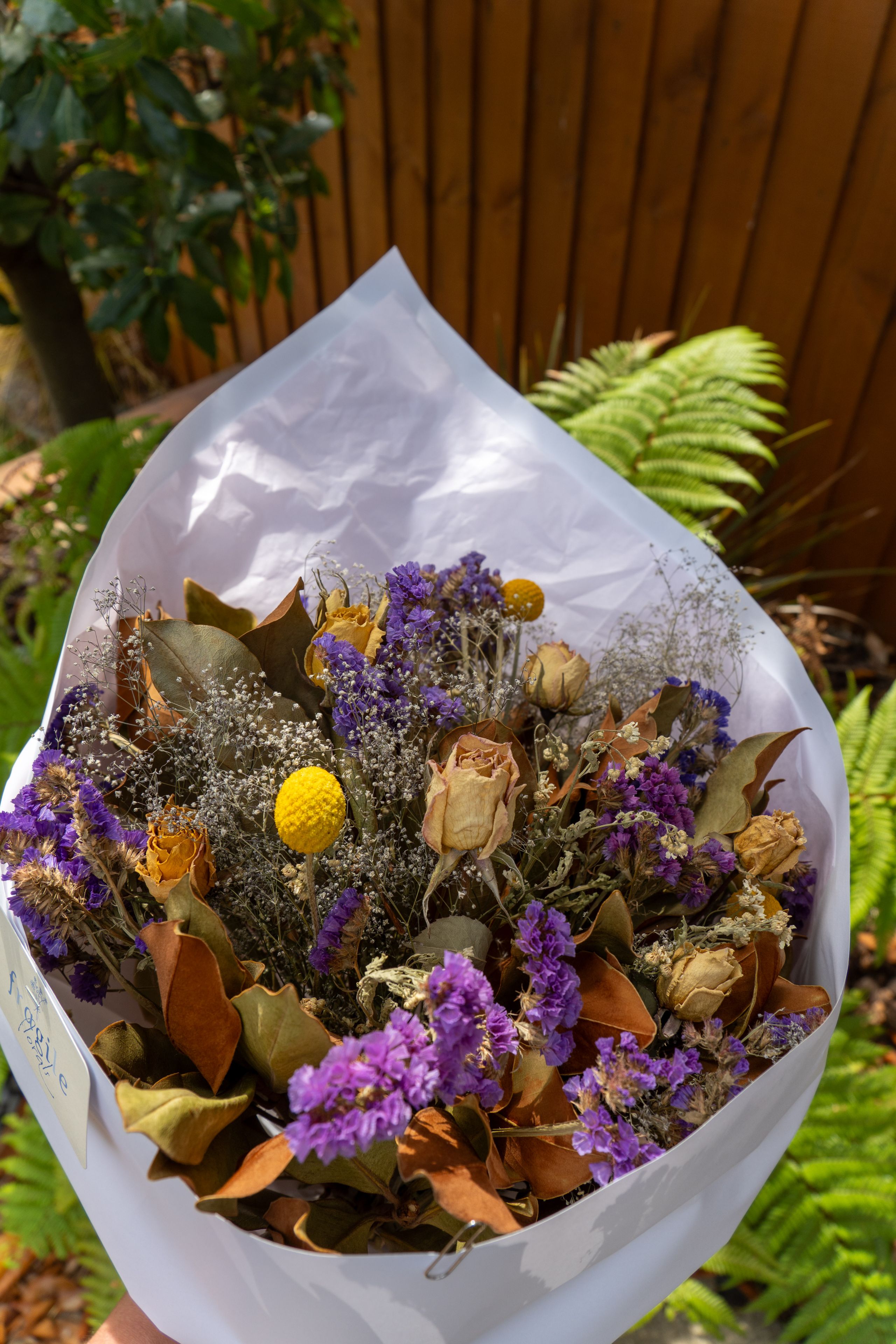 bouquet flowers dried purple yellow white leaves stems paper wrapping greenery background brown fence green ferns sunlight outdoor nature arrangement petals blossoms rustic decorative floral gift natural beauty garden romantic elegant vintage textured botanical composition vibrant colorful delicate artistic charming aesthetic fresh preserved ornamental handcrafted eco-friendly sustainable timeless classic serene peaceful harmonious tranquil lush foliage contrast bright soft subtle graceful enchanting lovely quaint picturesque quaint charming