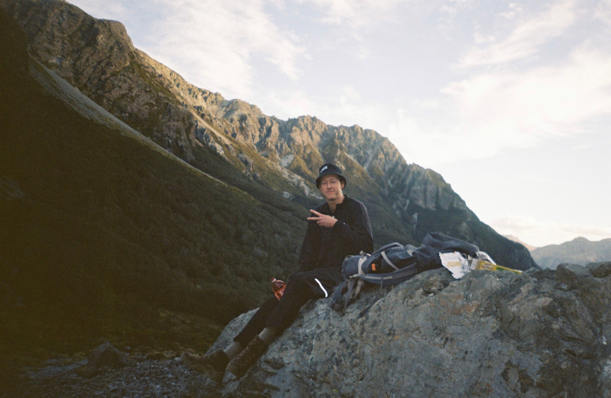 mountain rock person sky clouds jacket backpack hat sitting outdoor nature landscape hiking climbing blue gray green sunlight shadow rugged scenery adventure exploration travel wilderness