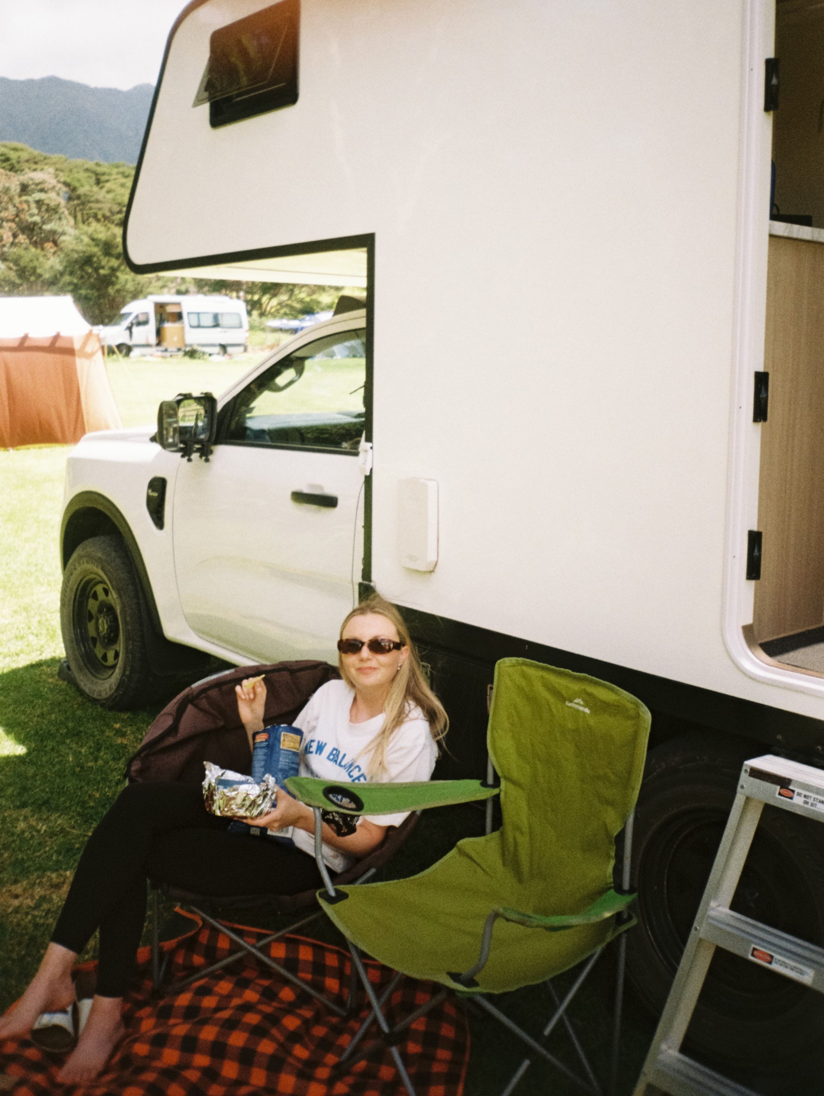 camper van woman sunglasses chair blanket ladder grass outdoors camping food drink vehicle white green black red sky trees mountain