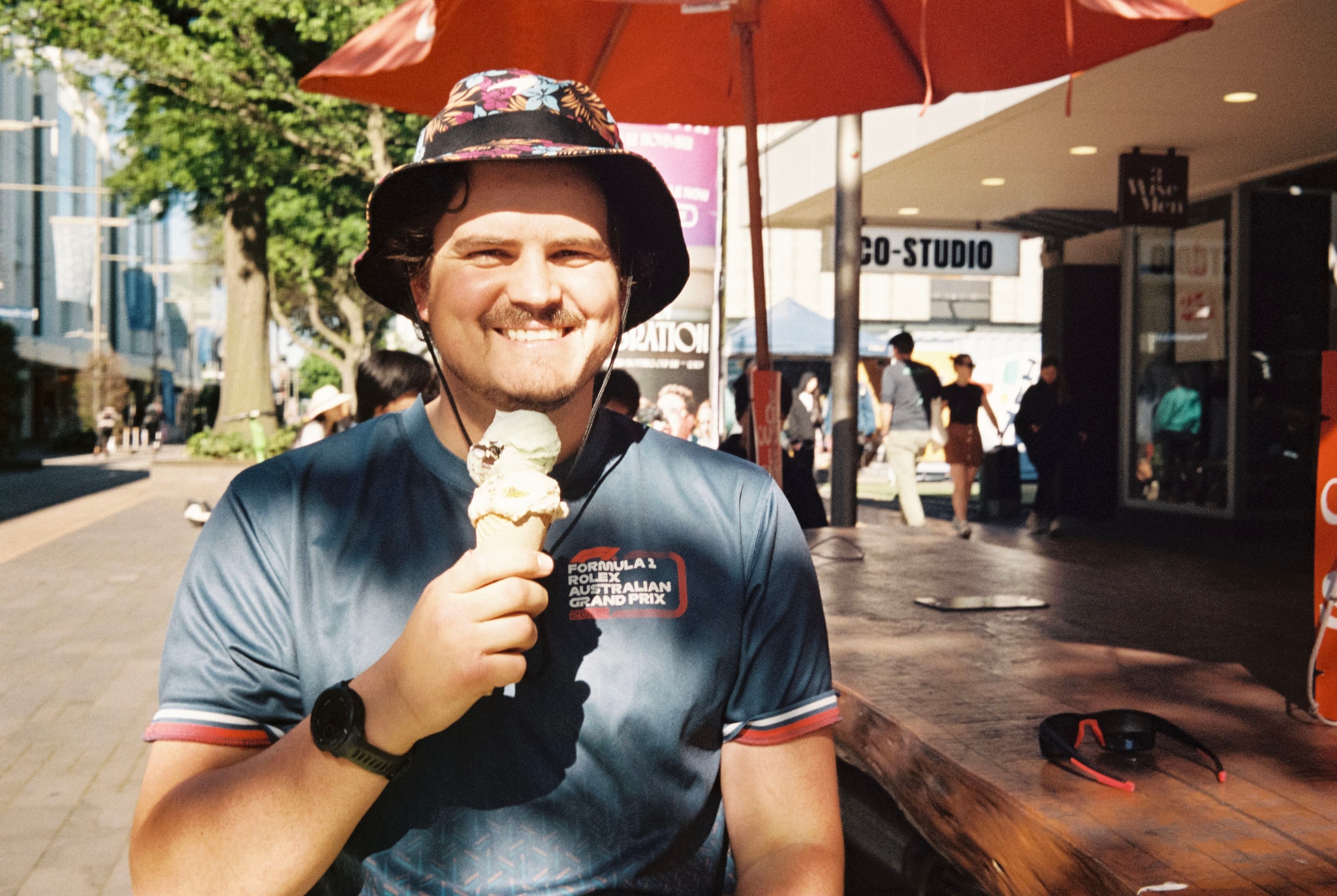 man hat ice cream cone t-shirt sunglasses wristwatch bench umbrella outdoor street building tree shadow sunlight smile summer casual red blue green brown white black people sidewalk shop sign day