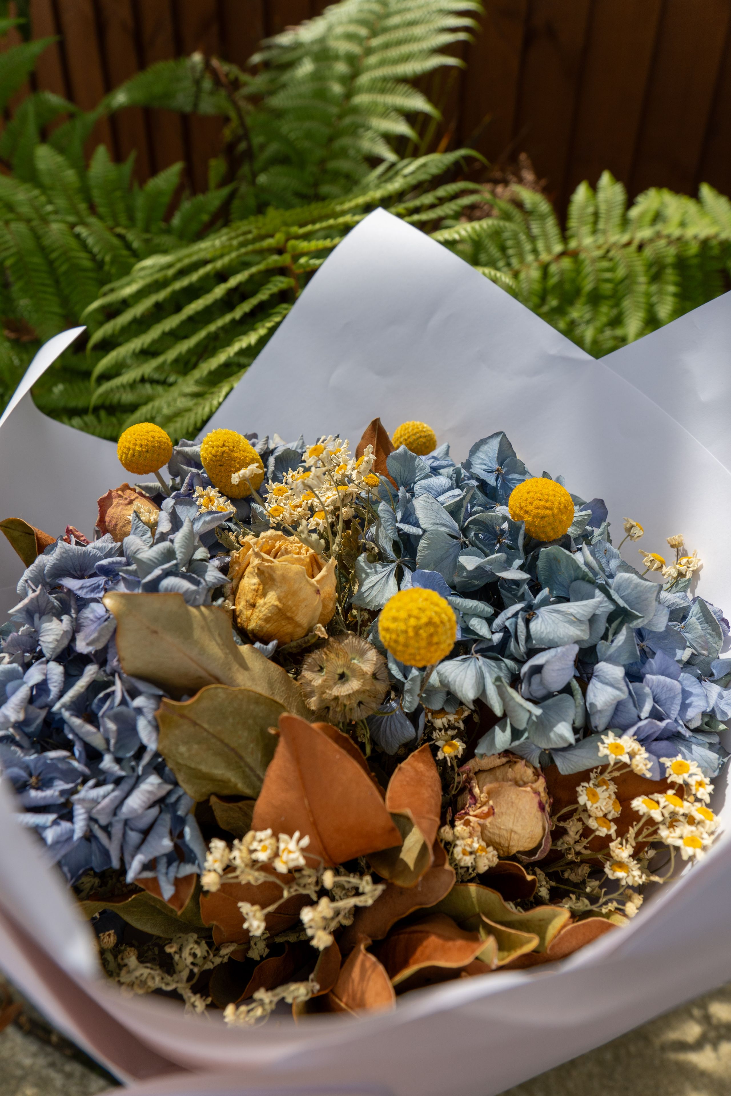 flowers bouquet hydrangea craspedia leaves foliage paper wrapping blue yellow brown green white garden outdoor nature arrangement blossoms petals stems ferns sunlight shadows texture natural rustic decorative floral vibrant colorful