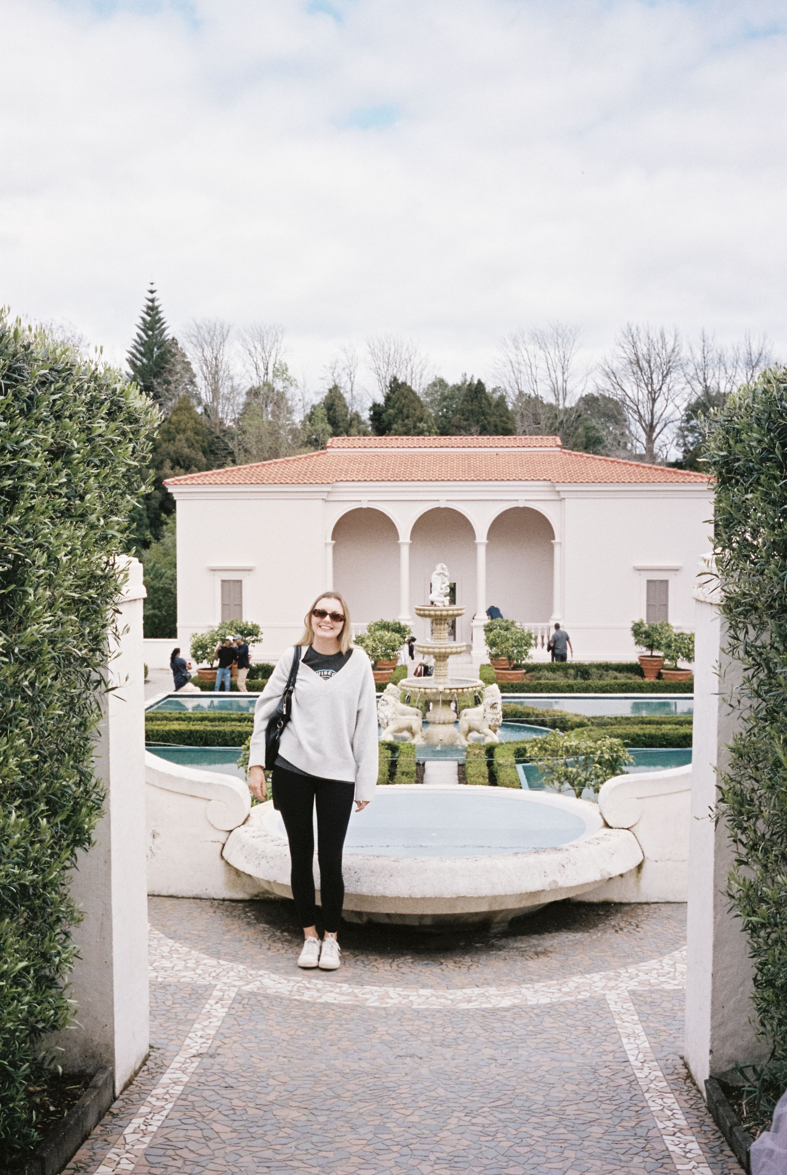 person garden fountain building arch trees sky plants pathway sunglasses sweater pants shoes bag white green blue red stone water architecture bushes walkway