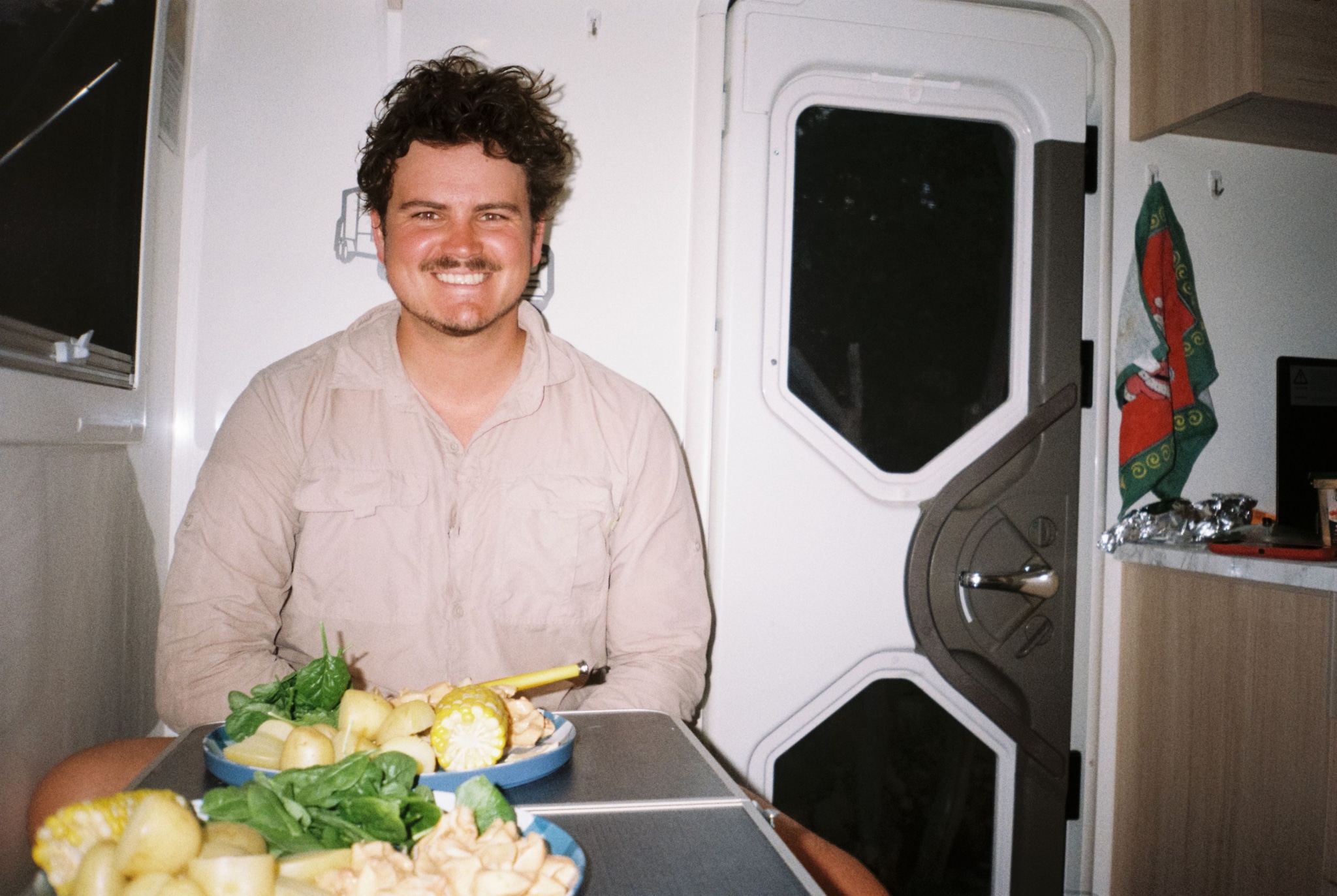 person smile table food vegetables potatoes cauliflower greens plate door interior flag red shirt beige