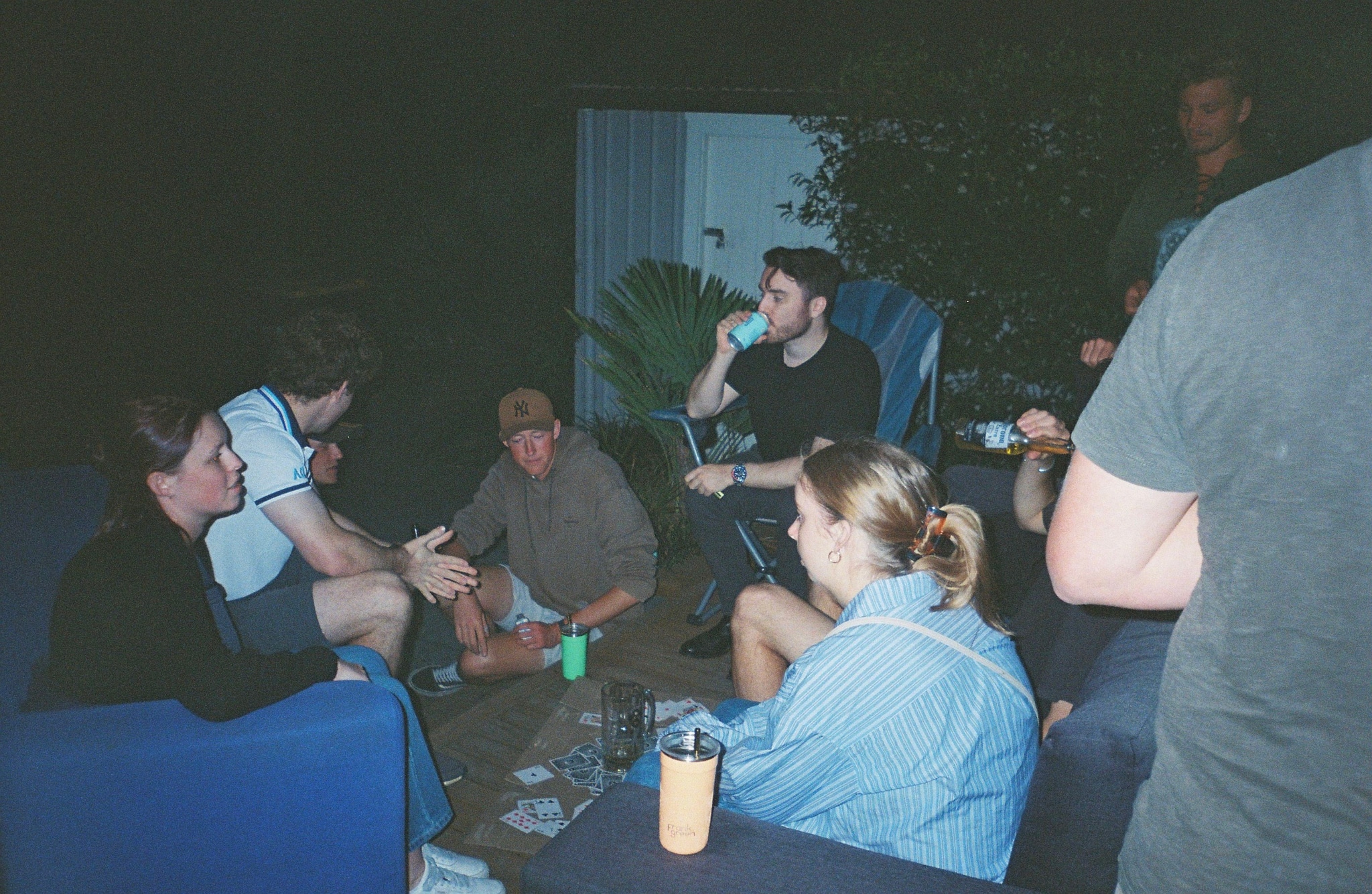 people group sitting talking drinking night outdoor chairs table plants blue black jacket shirt shorts can bottle cup conversation gathering socializing relaxation casual atmosphere