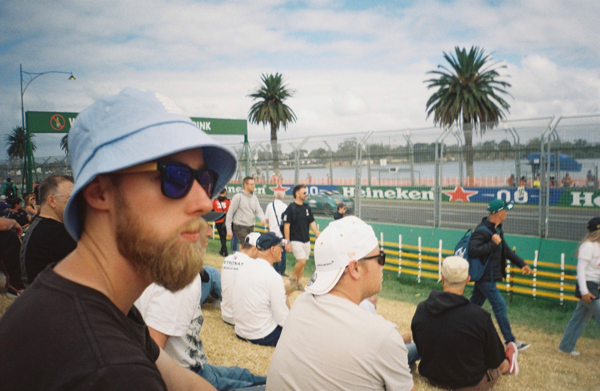 spectators hats sunglasses palm trees fence race track grass sky clouds blue people sitting standing outdoor event